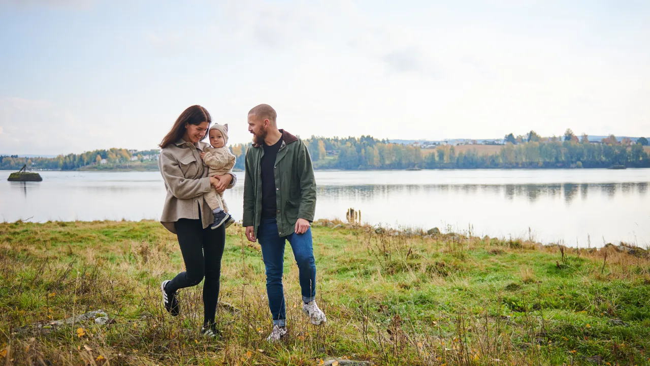 En mann og en kvinne holder en baby på et jorde ved en innsjø.