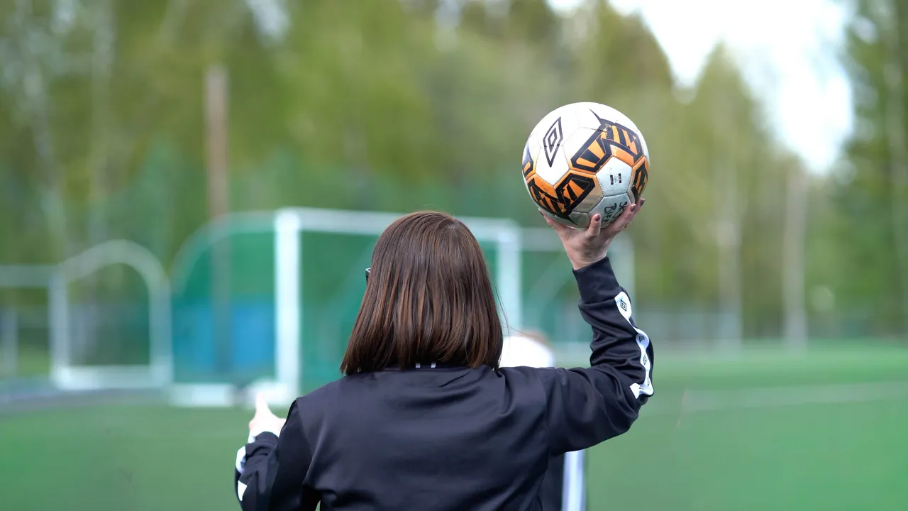 En kvinne, som står på en kunstgressbane, holder en ball i hånden og peker foran seg.