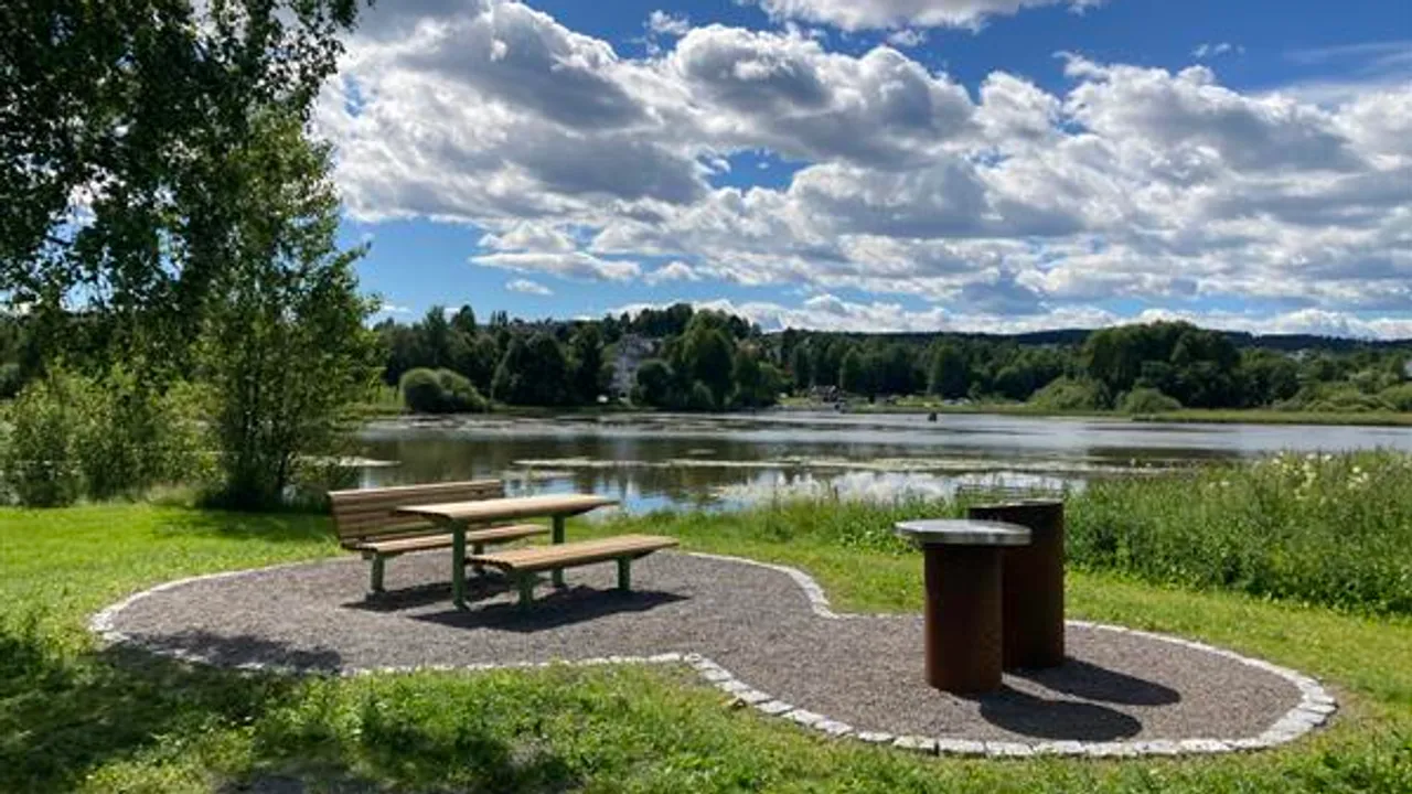 En park med en benk og en grill. Vann i bakgrunnen.