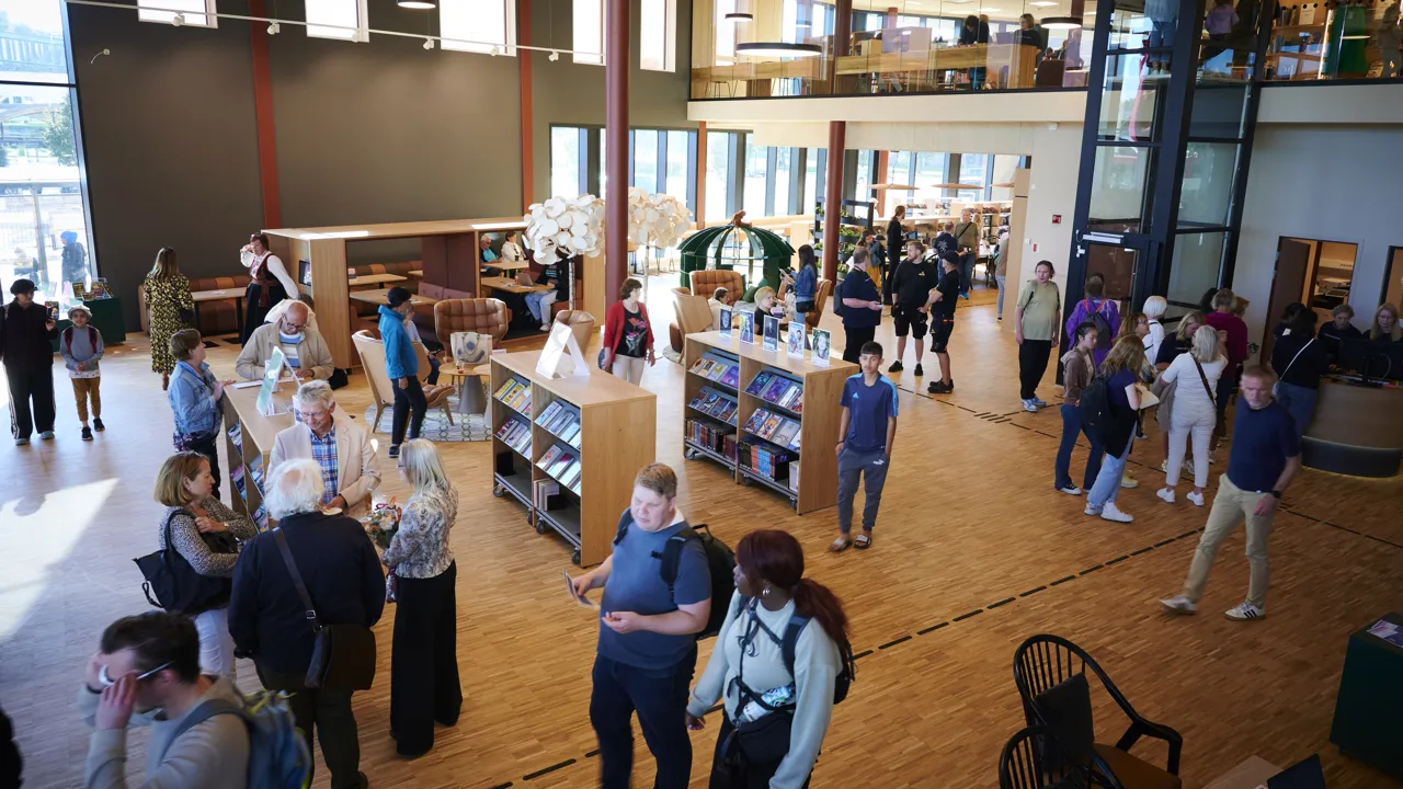 Mange mennesker går rundt eller står og snakker inne i et fellesområde på et bibliotek