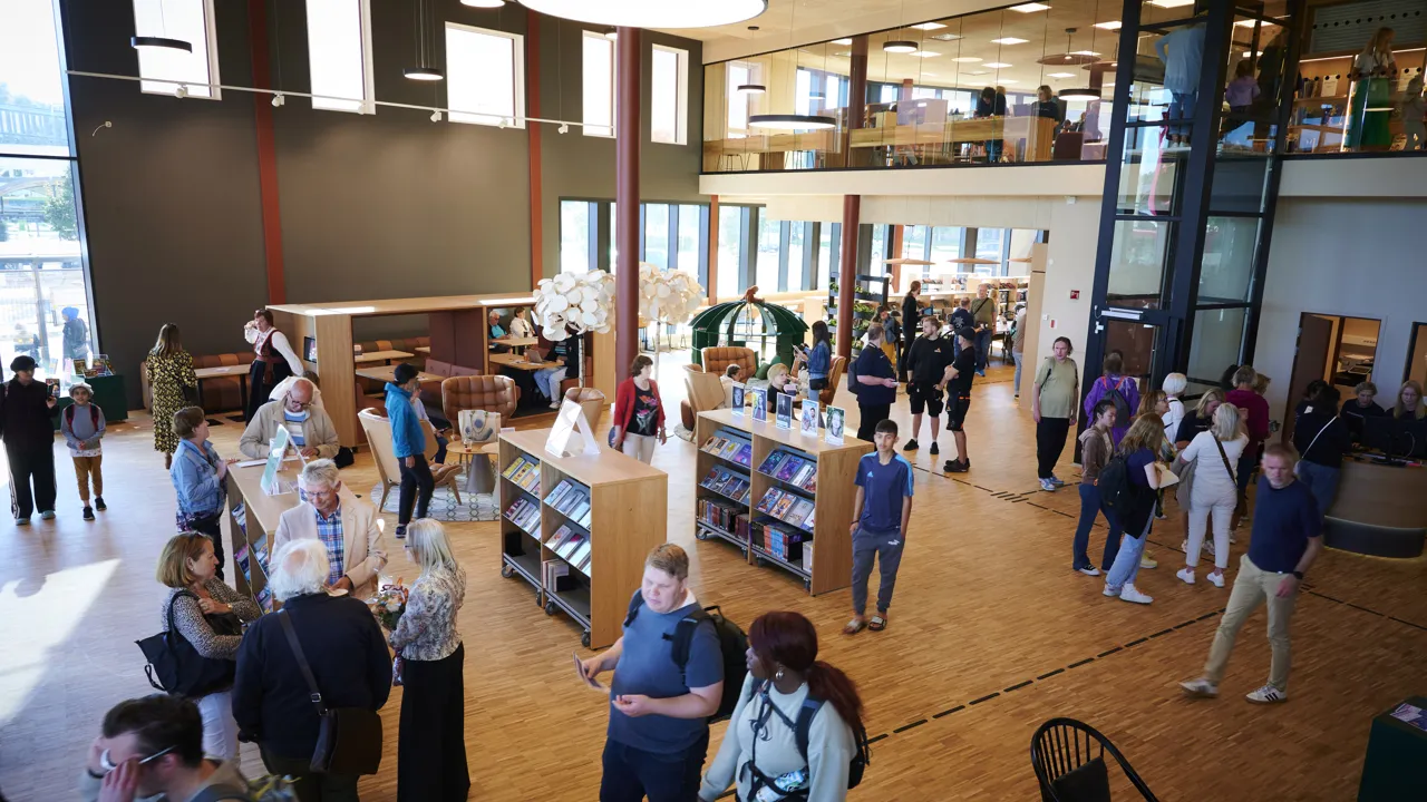 Mange mennesker går rundt eller står og snakker inne i et fellesområde på et bibliotek