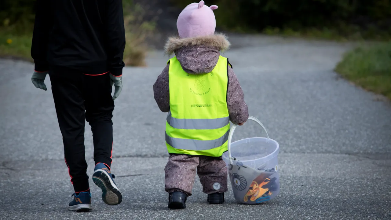 To barn går langs en vei. Den minste har på seg refleksvest og bærer en bøtte med søppel