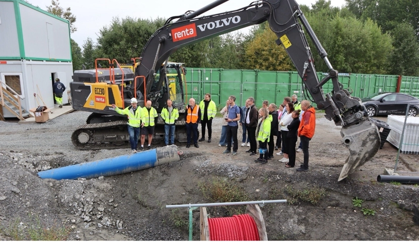 Anleggsarbeidere som står foran en gravemaskin.