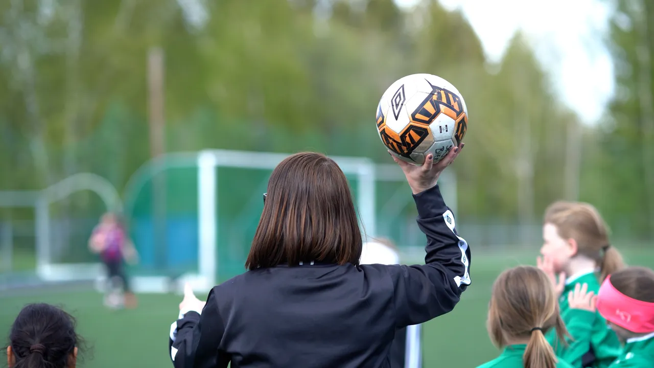 En kvinne holder en fotball foran en gruppe barn.